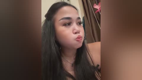 Media: A video of a young Asian woman with long black hair, light skin, and full lips, blowing a kiss, in a dimly lit room with a brown curtain and pink decorations.