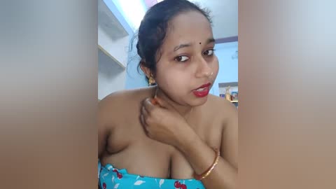 Media: A video of a South Asian woman with medium brown skin, dark hair, and red lipstick, wearing a blue strapless dress with a floral pattern. She stands indoors, in front of a blurred background.