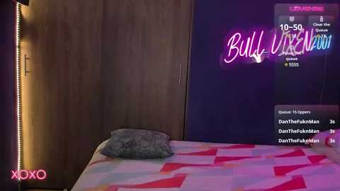 Media: Video of a bedroom with a pink and white patterned bedspread, a gray pillow, a wooden wardrobe, and a neon \"Bullseye\" sign on the wall.