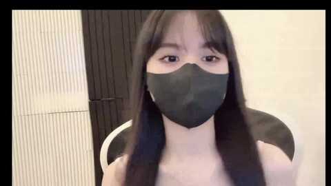 Media: Video of an Asian woman with long black hair, wearing a black face mask, sitting indoors. She has a pale complexion, and the background features a beige wall and a wooden door.