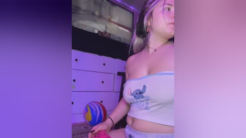 Media: Video of a young woman with blonde hair, wearing a white crop top and denim shorts, holding a colorful ball, in a dimly lit room with purple lighting.