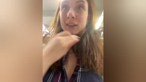 Media: Video of a young woman with fair skin and long brown hair, wearing a plaid shirt, smiling while adjusting her hair. Background shows blurred bookshelves and lighting.