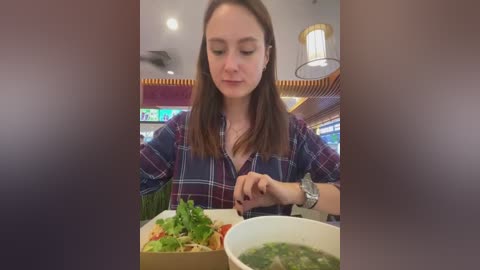 Media: A video of a young woman with straight, brown hair, wearing a plaid shirt, eating soup at a restaurant with a modern interior and neon signs in the background.