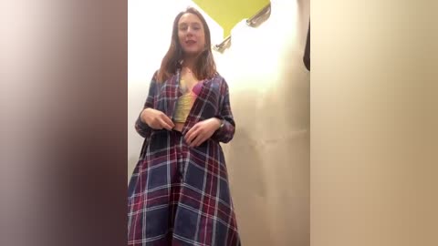 Media: Video of a woman with long brown hair, wearing a plaid robe, standing in a beige-walled, dimly lit bathroom with a yellow ceiling.