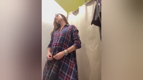 Media: Video of a woman in a plaid dress, standing in a fitting room with beige walls and a green ceiling. She has her eyes closed, holding the dress's hem, and there are hanging clothes on a rack in the background.