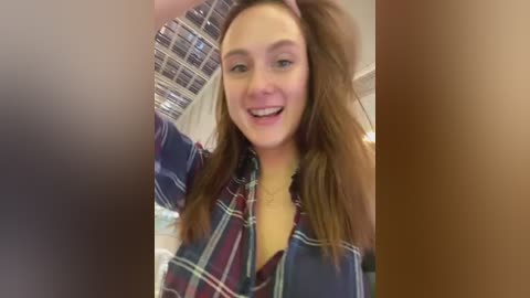 Media: Video of a young woman with light skin and long brown hair, smiling and holding her hair. She wears a blue and red plaid shirt. Background shows a tiled wall and blurry figures.