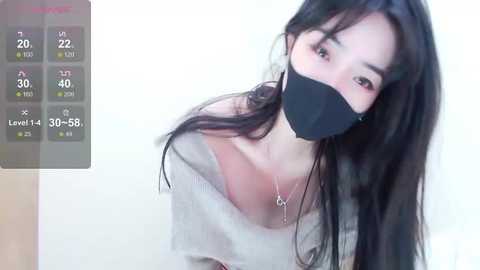 Media: A video of an Asian woman with long black hair, wearing a black face mask and a beige off-shoulder top, against a plain white background.