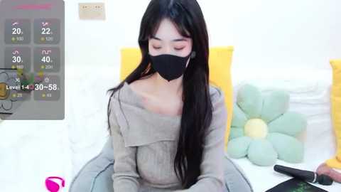 Media: A young Asian woman with long black hair, wearing a black face mask, sits in a bright room with a yellow bed and a green flower pillow.