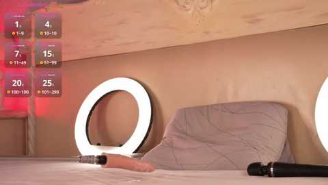 Media: A video of a cozy bedroom with a beige wall and a white bed, featuring a pillow with a quilted pattern and a pink dildo on the bed. A ring light and a black dildo are on the bedside table.