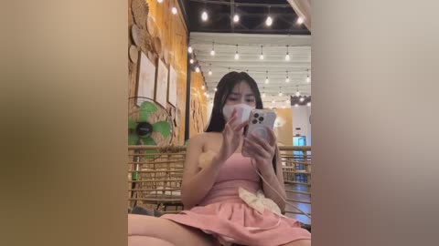 Media: Video of an Asian woman with shoulder-length black hair, wearing a pink dress, sitting on a wooden bench, taking a selfie in a rustic, warmly lit cafe with string lights, wicker chairs, and a green fan.