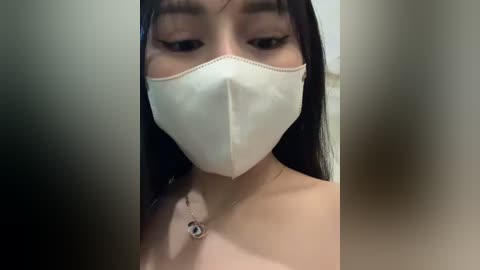 Media: Video of an Asian woman with long black hair and light skin, wearing a white surgical mask and a delicate necklace. The background is blurred, focusing attention on her face.