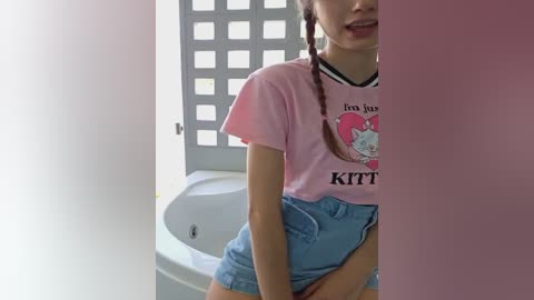 Media: A video of a young woman with light skin and long red braids, wearing a pink t-shirt with a cat graphic and blue denim shorts, standing in a bathroom with a white sink and frosted glass window.