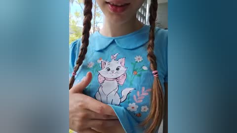 Media: Video of a young girl with light skin and long brown braided hair, wearing a blue collared shirt with a cartoon cat design. She's indoors with a blurred background.