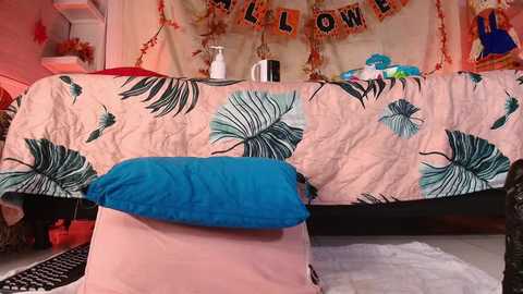 Media: Video of a cozy bedroom with a bed draped in a pink, palm leaf-patterned blanket and a blue pillow. Behind, a \"Welcome\" banner and festive decor adorn the walls.