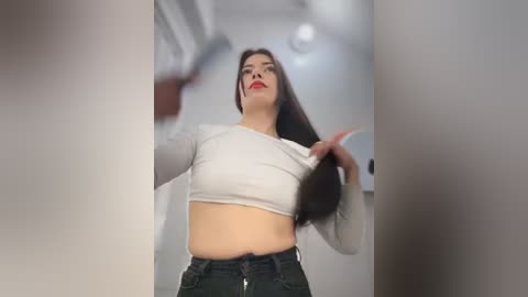 Media: Video of a curvy woman with long dark hair, wearing a white cropped top and black jeans, holding her hair. She stands in a brightly lit, modern room with white walls and a ceiling light.