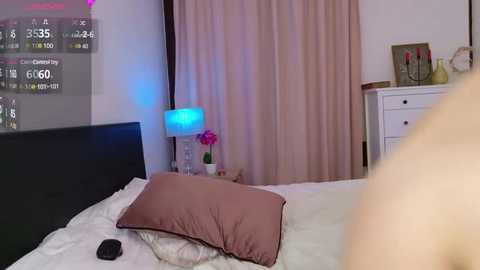 Media: Video of a cozy bedroom with a black headboard, pink pillow, white sheets, white dresser, pink curtains, and a glowing blue lamp on a nightstand.