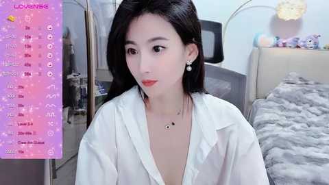 Media: A video of an Asian woman with long black hair, wearing a white blouse, in a bedroom with a gray bed and stuffed toys.