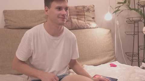 Media: Video of a young man with short brown hair, wearing a white t-shirt and denim shorts, sitting on a beige sofa in a cozy living room with a potted plant and a round metal table lamp.