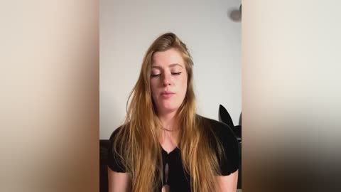 Media: A video of a young Caucasian woman with long, straight blonde hair, wearing a black top, standing in a dimly lit room with beige walls, her eyes closed in a serene expression.