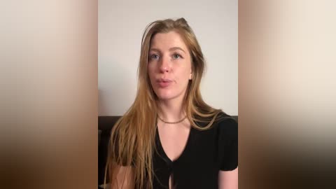 Media: A video of a young woman with long, straight, light brown hair, fair skin, and a slender physique, wearing a black V-neck top. She's indoors, with a neutral background and minimalistic setting.