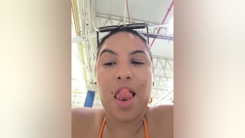 Media: Video of a young woman with medium skin tone, dark hair, and an orange bikini, sticking out her tongue in a playful pose. Background shows an industrial setting with metal beams and pipes.