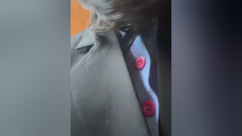 Media: Close-up video of a person's neck and shoulder, wearing a light gray suit with a white shirt and bright pink buttons. The background is blurred, with an orange wall on the left and a dark wall on the right.