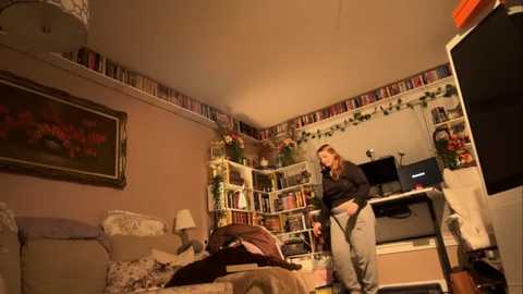 Media: Video of a cozy, warmly-lit bedroom with a woman in a black sweater and gray pants playing a video game on a TV stand. Bookshelves and floral decor adorn the walls.