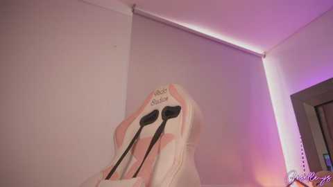 Media: A video of a pink and white gaming chair with black headphones and a white pillow, set against a beige wall with purple lighting, creating a cozy, modern gaming setup.