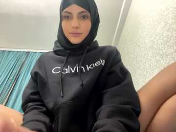 Media: Video of a young woman with olive skin, wearing a black Calvin Klein hoodie, black hijab, and a contemplative expression. Background features teal curtains and textured wallpaper.