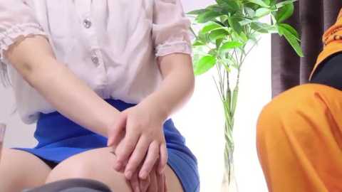 Media: Video of a light-skinned woman with a white blouse and blue skirt massaging a man's back, who is wearing an orange shirt, in a brightly lit room with a potted plant in the background.