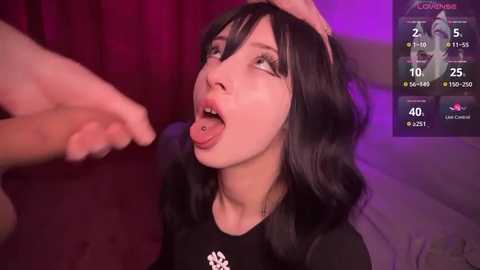 Media: Video of a young woman with fair skin and long black hair, wearing a black shirt, sticking her tongue out while a hand touches her head. Background shows a dimly lit room with purple lighting and a digital clock.