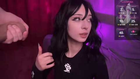 Media: Video of a pale-skinned woman with long black hair and makeup, giving a thumbs-up in a dimly-lit room, with a digital overlay showing a game score and stats.