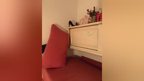 Media: A video of a cozy, dimly lit room with a cream-colored wooden dresser topped with a bouquet of pink and red flowers. A red pillow rests on a red bedspread. The background is a warm, reddish-orange wall.