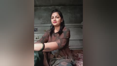 Media: Video of a South Asian woman with dark hair, wearing a brown patterned sari, sitting on a bed in a dimly lit room with gray walls and a wooden shelf.