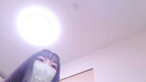 Media: Video of a young woman with long, dark hair, wearing a light blue face mask, standing in a brightly lit room with a circular light fixture.