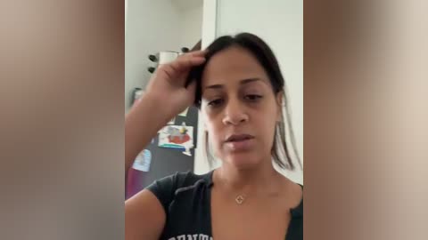 Media: Video of a woman with medium brown skin and long black hair, wearing a black t-shirt, standing in a room with a white door and a colorful poster in the background.