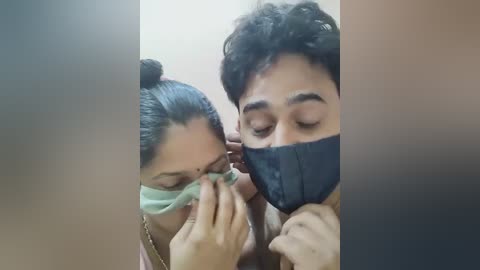 Media: Video of a man and woman, both with masks on, the woman applying a face mask on the man. The background is blurred.