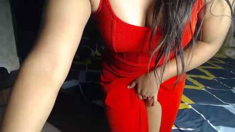 Media: Video of a curvy woman with long dark hair in a bright red, low-cut dress, standing indoors with a patterned bedspread in the background.