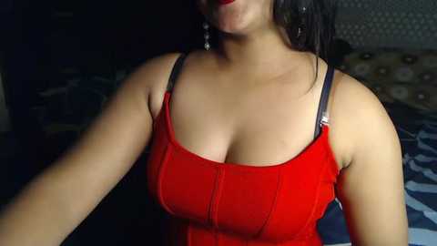 Media: Video of a woman with medium skin tone, wearing a red corset top, black straps, and dangling earrings. Background shows dimly lit bedroom with a patterned headboard and a bed with blue and white bedding.