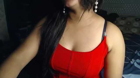 Media: Video of a woman with long black hair wearing a red tank top, silver earrings, and a subtle patterned background.