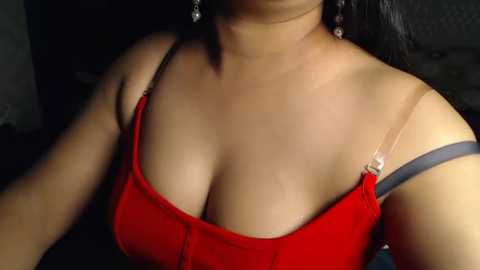 Media: Video of a woman with medium brown skin, wearing a red tank top with thin straps, showcasing her ample cleavage. She has long black hair, and her face is partially visible, adorned with dangling earrings.