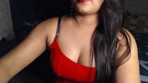 Media: A video of a South Asian woman with long, dark hair, wearing a red, low-cut, sleeveless top, sitting in a dimly-lit room. Her full lips are slightly parted, revealing a hint of her teeth.