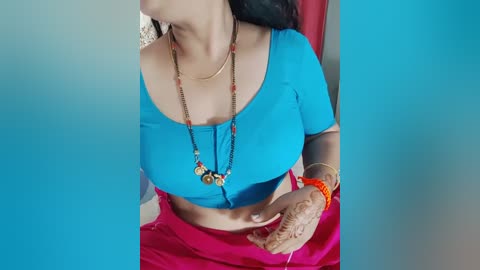 Media: A video of a woman in a blue crop top, red skirt, and intricate henna designs on her hands. She wears a long, beaded necklace and an orange bracelet. The background is a gradient of blue and red.