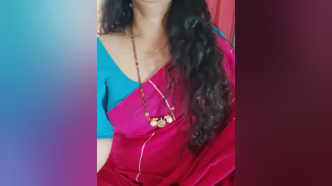 Media: A video of a woman with long, wavy black hair, wearing a vibrant blue and magenta sari with a gold necklace. The background is a gradient of blue to red.