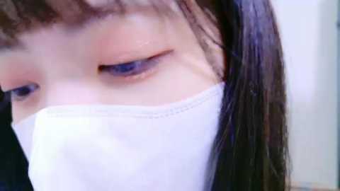 Media: Video of a person with light skin, long dark hair, wearing a white mask, with blue eyeshadow and light makeup, close-up of the face, blurry background.