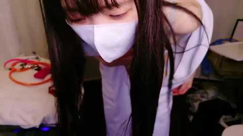 Media: Video of an Asian woman with long black hair, wearing a white surgical mask, bending forward, in a cluttered room with a bed, orange cords, and a desk.