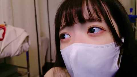 Media: A video of a young Asian girl with dark hair and blue eyes, wearing a white surgical mask, standing in a medical setting with white curtains and a folded white gown on a chair.