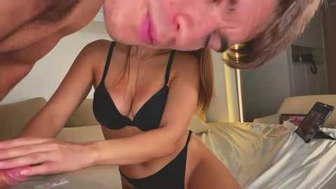 Media: Video of a woman with light skin and blonde hair, wearing a black bra and panties, lying on a bed with a man's face in close proximity.