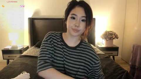 Media: Video of a young Asian woman with dark hair, wearing a striped shirt, smiling, in a cozy bedroom with a floral arrangement on a nightstand.