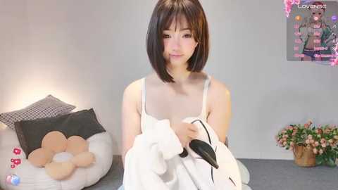 Media: A video of a young East Asian woman with a straight, shoulder-length bob haircut, wearing a white spaghetti-strap tank top, sitting on a bed. She holds a black smartphone.